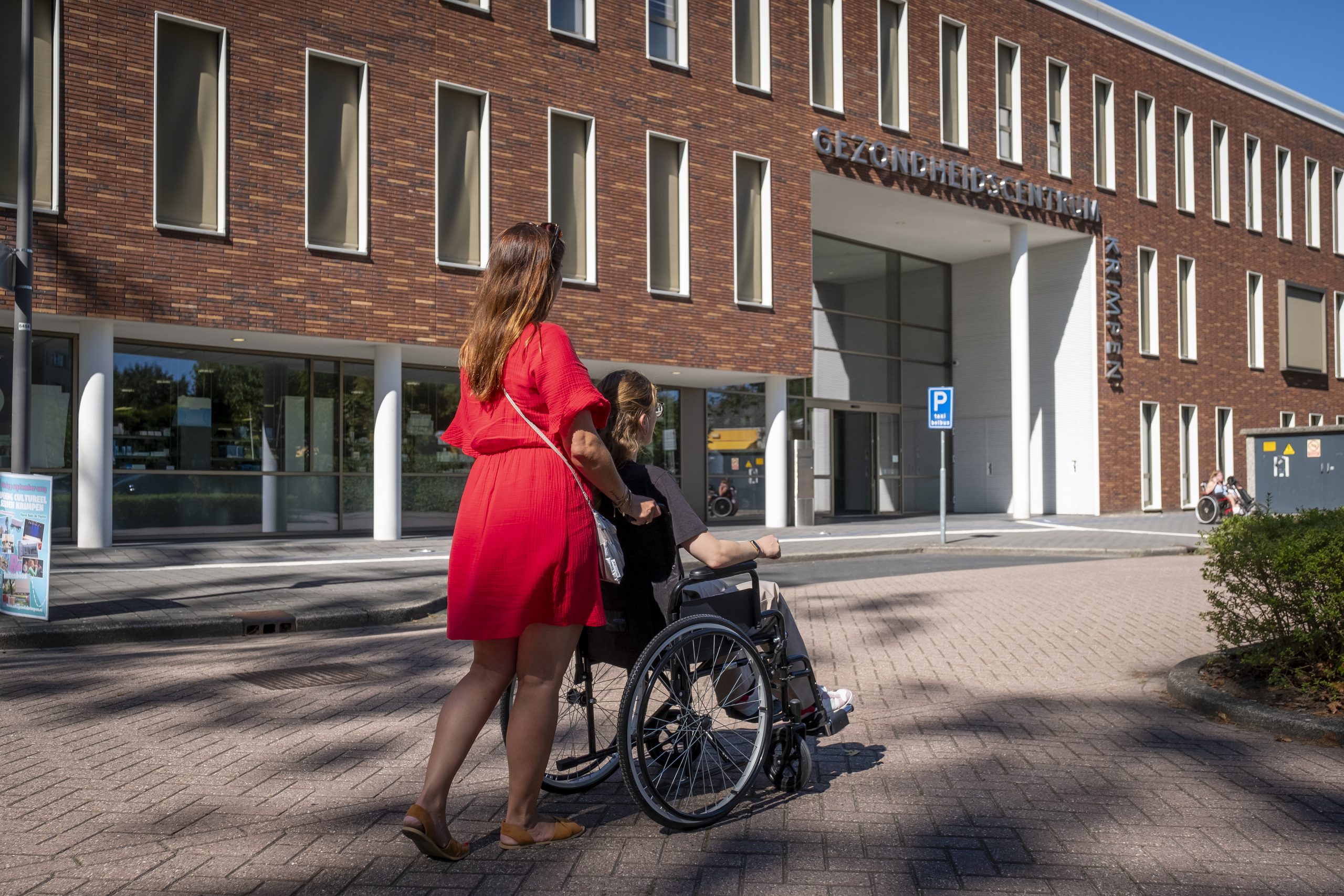Afbeelding van het Gezondheidscentrum, met op de voorgrond 2 personen waarvan 1 in een rolstoel.