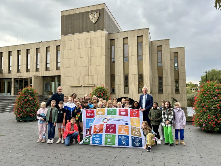 Leerlingen uit groep 6 poseren samen met wethouder Wubbo Tempel voor het raadhuis met de Global Goals Day-vlag